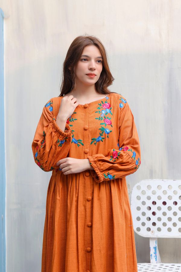 Woman wearing an orange dress with floral embroidery against a light curtain background