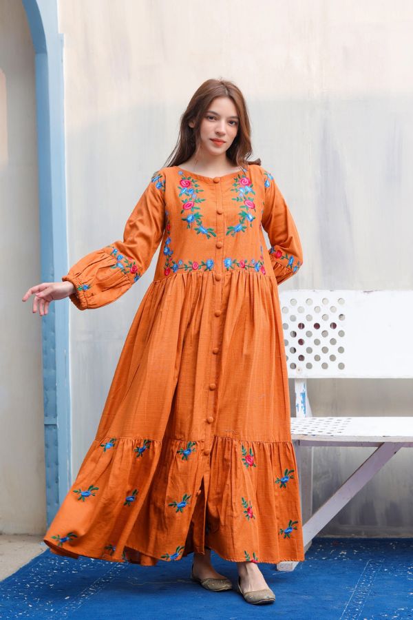Woman wearing an orange dress with floral embroidery standing against a light-colored wall.