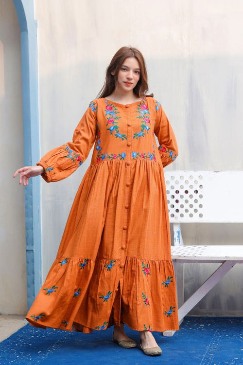 Woman wearing an orange dress with floral embroidery standing against a light-colored wall.