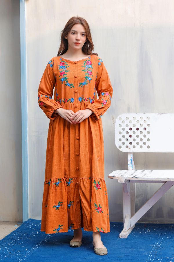 Woman wearing an orange dress with floral embroidery standing in a room with a white chair and blue floor.
