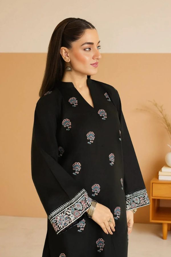 Woman wearing a black traditional outfit with intricate patterns in a room with beige walls and a wooden side table.