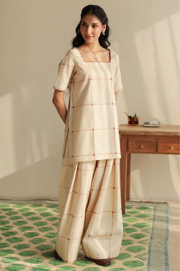 Woman wearing a traditional outfit with a checkered pattern in a room with a wooden desk and green patterned rug.