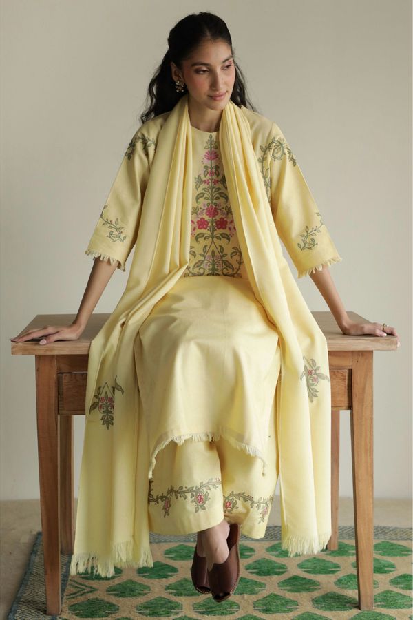 Woman in a light yellow traditional outfit with floral patterns sitting on a wooden table.