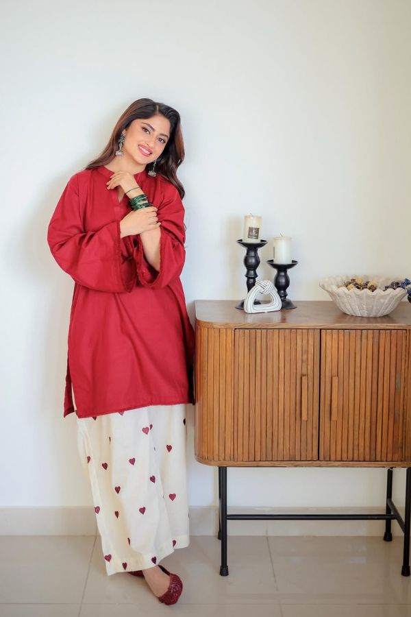 Woman in a red top and white pants with heart patterns standing next to a wooden cabinet.