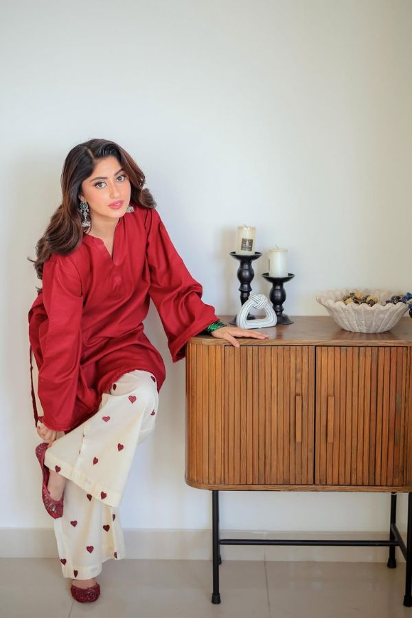 Woman in a red top and white pants with heart patterns leaning against a wooden cabinet.