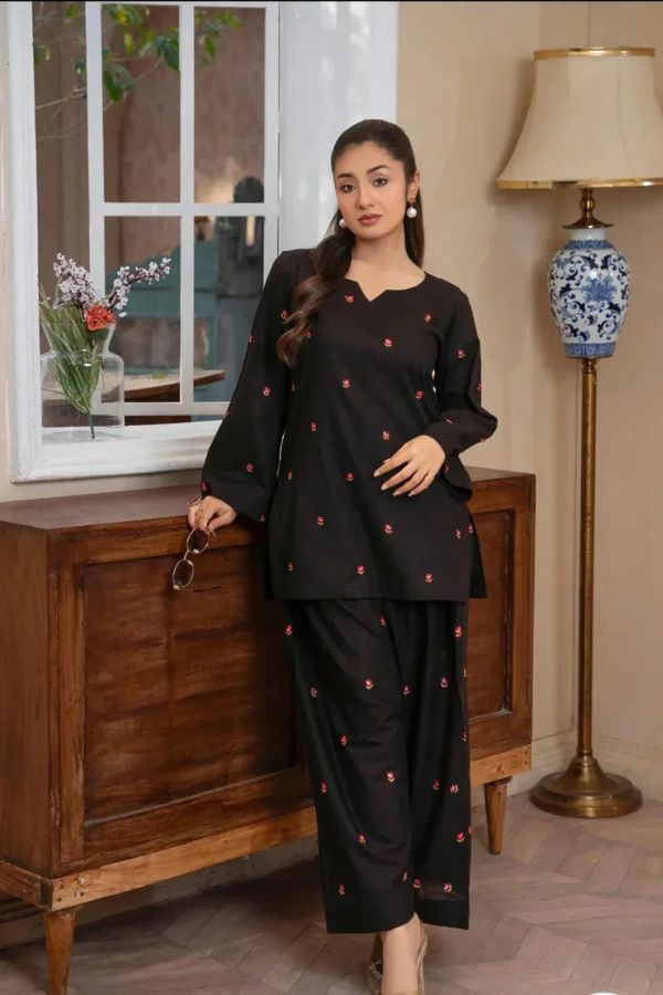 Woman in a black outfit with red patterns standing in a room with wooden furniture and a lamp.