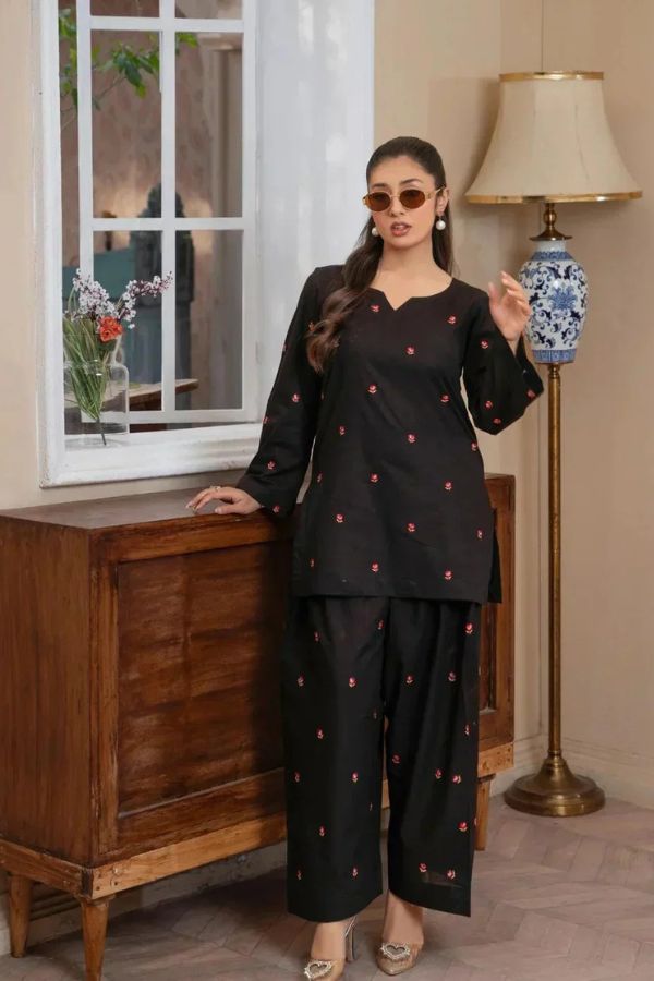 Woman in a black outfit with red patterns standing in a room with a lamp and window.