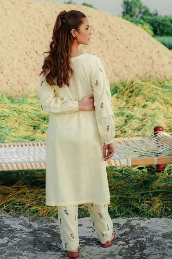 Woman in a light-colored traditional outfit standing in a rural setting with a haystack and greenery.