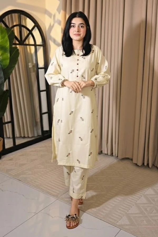 Woman in a cream-colored outfit standing indoors with curtains and a plant in the background