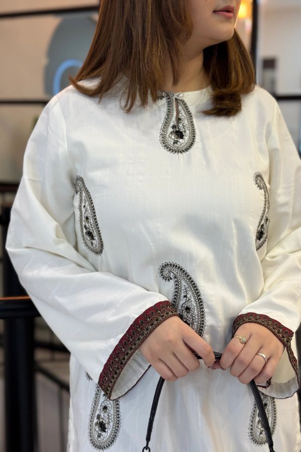 Person wearing a white outfit with intricate black embroidery in a blurred indoor setting