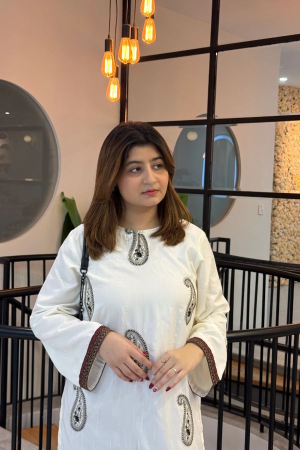 Woman in a white traditional outfit with intricate patterns in an indoor setting.