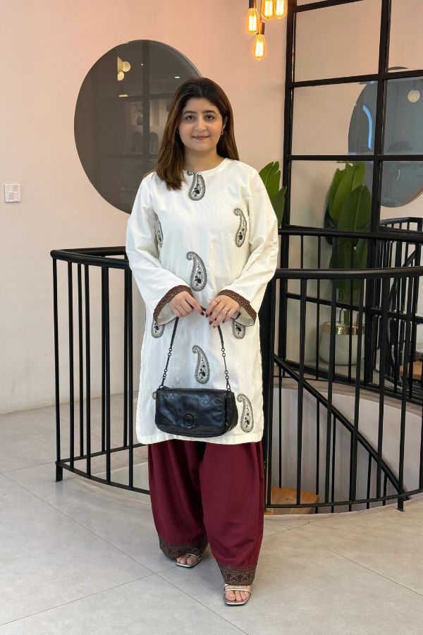 Woman in a white traditional outfit with maroon pants holding a black handbag indoors.