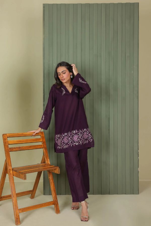Woman in a purple embroidered outfit standing next to a wooden chair against a green striped backdrop.