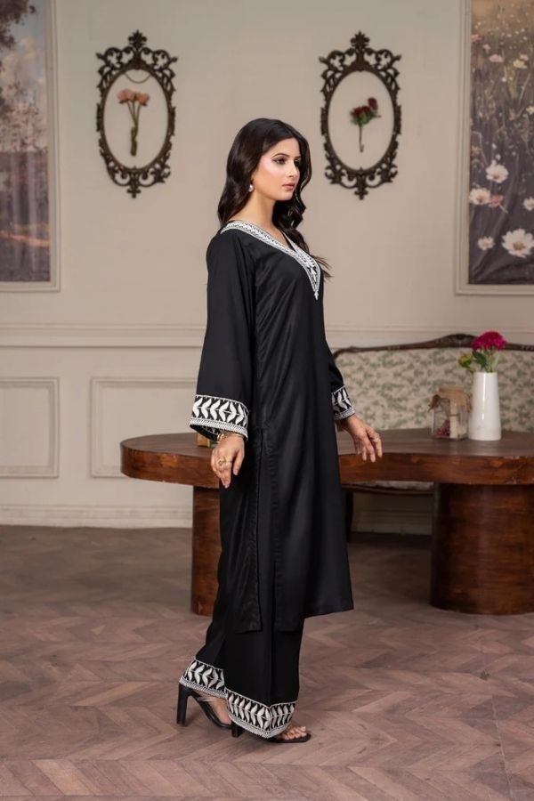 Woman in a black dress with white patterns standing in a room with a wooden table and decorative wall frames.