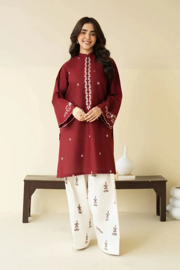 Woman wearing a maroon embroidered top and white pants against a beige background