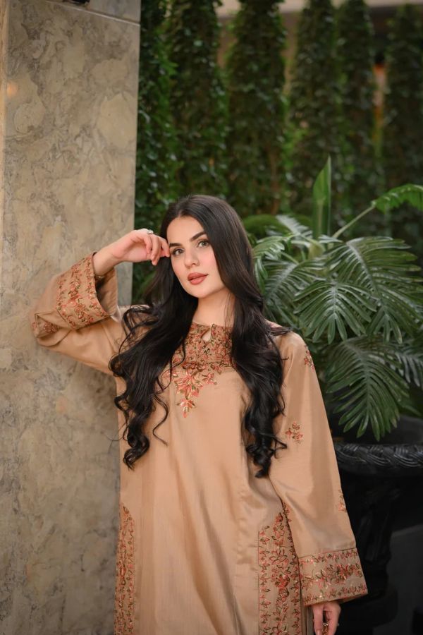 Woman wearing a beige embroidered outfit with long dark hair, standing against a green leafy background.