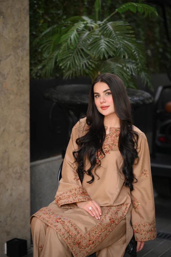 Woman in a beige embroidered outfit sitting outdoors with plants in the background