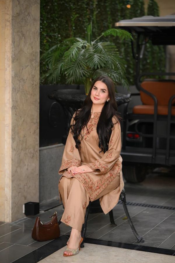 Woman in a beige floral dress sitting on a chair with plants and a vehicle in the background.