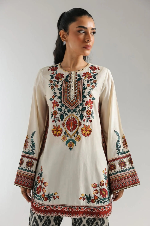 Woman wearing a cream-colored embroidered top with colorful patterns on a plain background