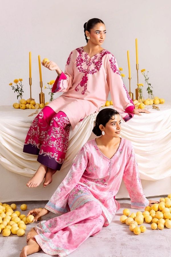 Two women in pink and purple traditional outfits sitting on a white draped surface with yellow flowers.