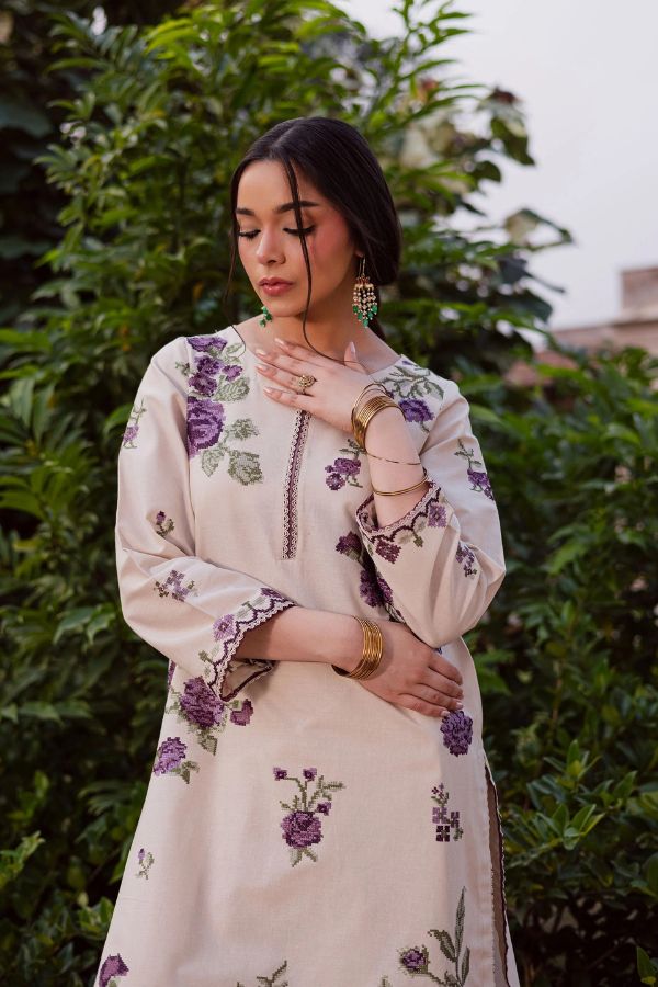 Woman wearing a cream-colored embroidered outfit with purple floral patterns against a green foliage background.