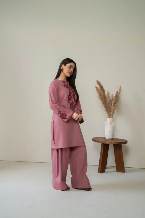 Woman in a pink outfit standing next to a wooden stool with a vase and dried plants against a plain background