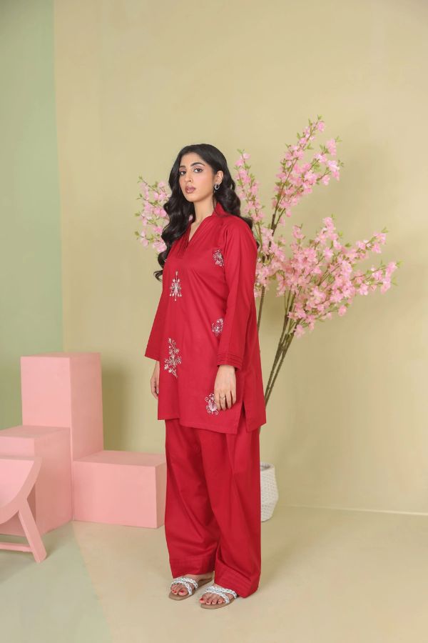 Woman in a red outfit with floral embroidery standing next to pink flowers against a light green wall.