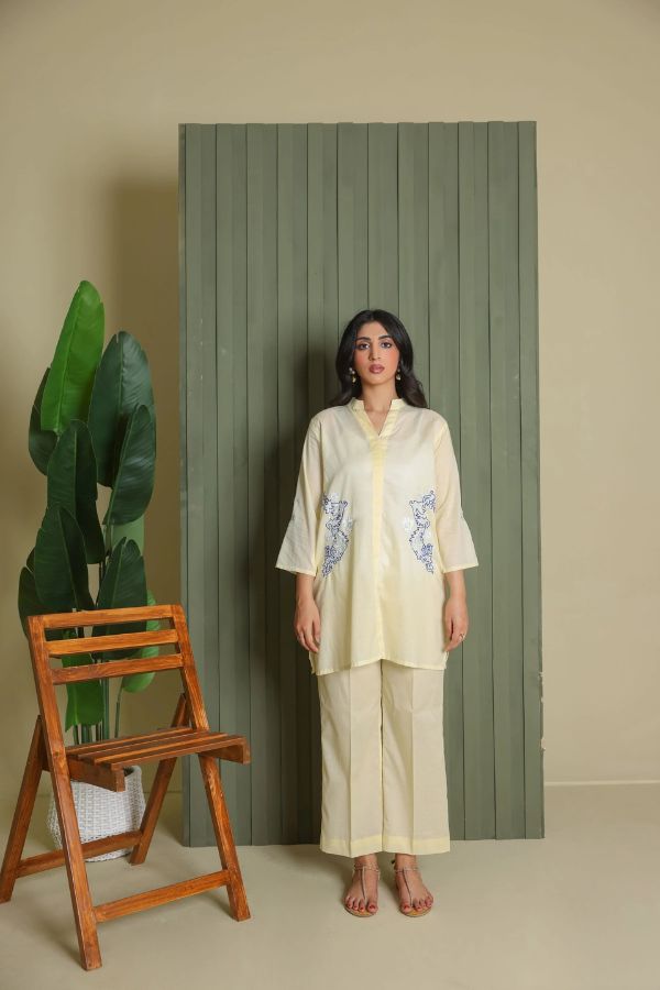 Woman in a white outfit standing next to a wooden chair and green plant against a gray wall.