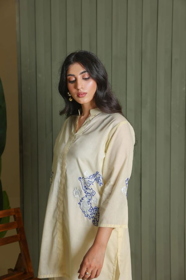 Woman wearing a light-colored outfit with blue embroidery against a green wall.