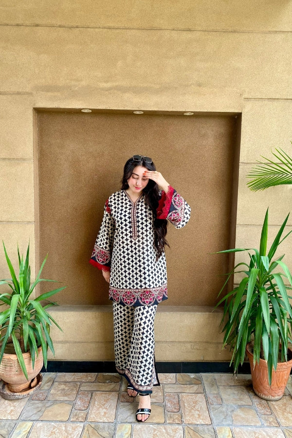 Woman in a patterned outfit standing in front of a beige wall with plants on either side.