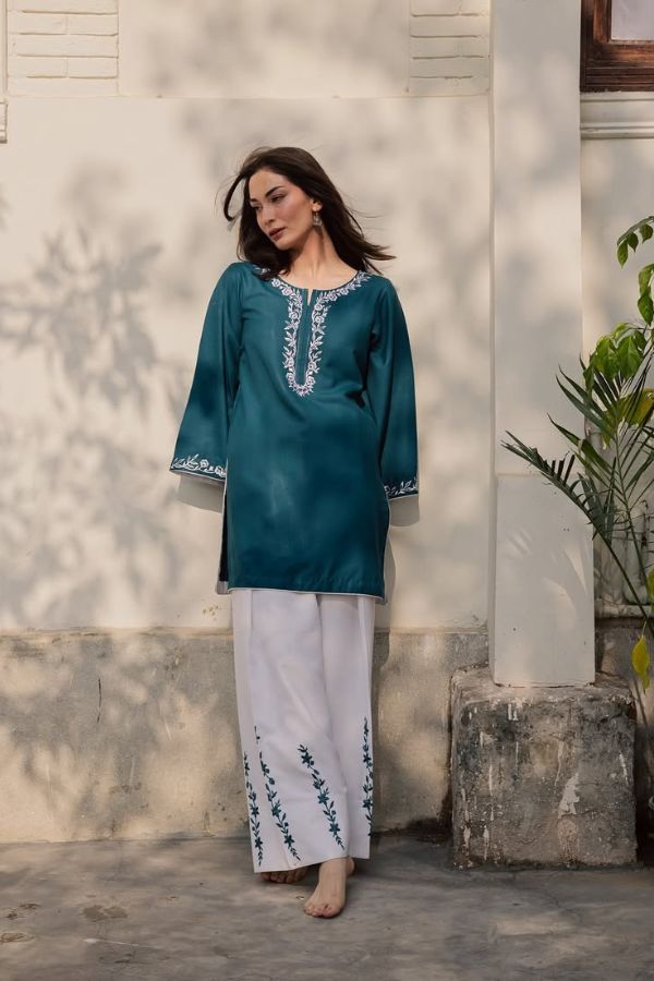 Woman wearing a teal embroidered top and white pants standing against a light-colored wall with a plant in the background.