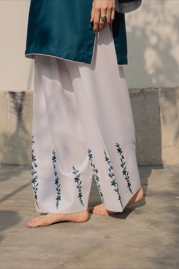 Person wearing white pants with floral embroidery and a teal top against a neutral background