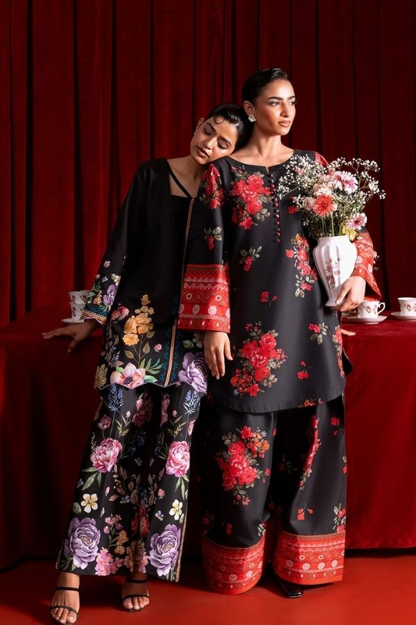 Two women in black floral outfits standing in front of a red curtain with a table set for tea.