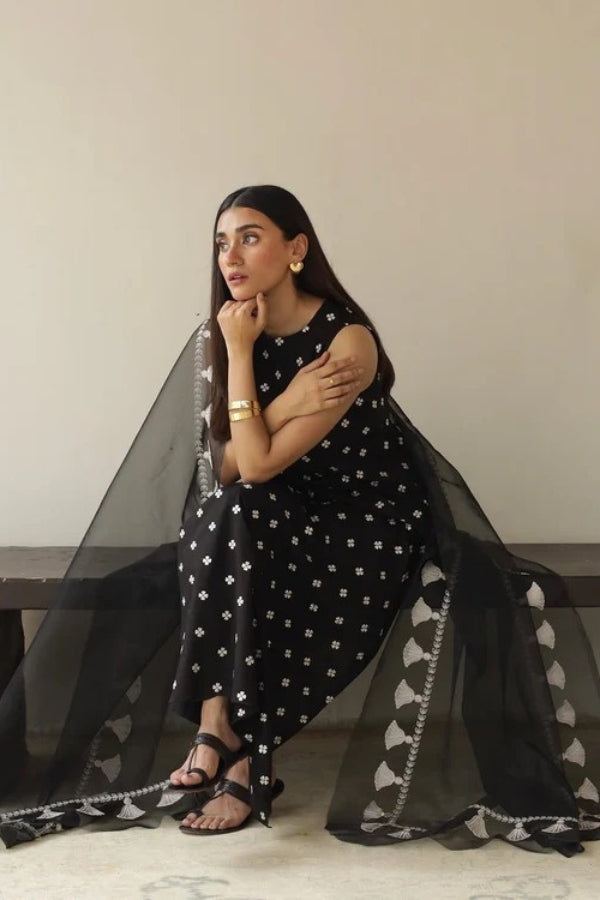 Woman in a black and white patterned outfit with sheer black fabric draped over her, sitting on a light-colored floor.