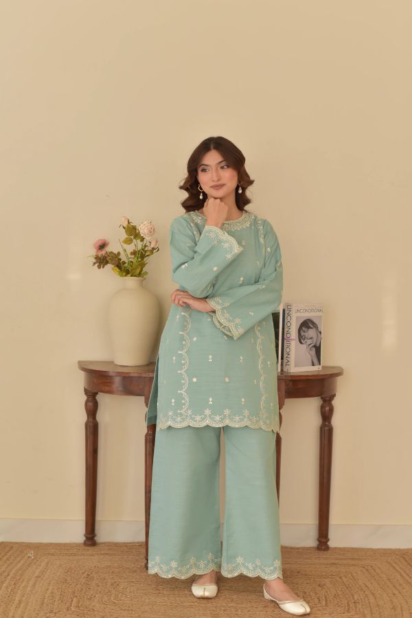 Woman in a light blue traditional outfit standing in a room with a table and vase in the background.