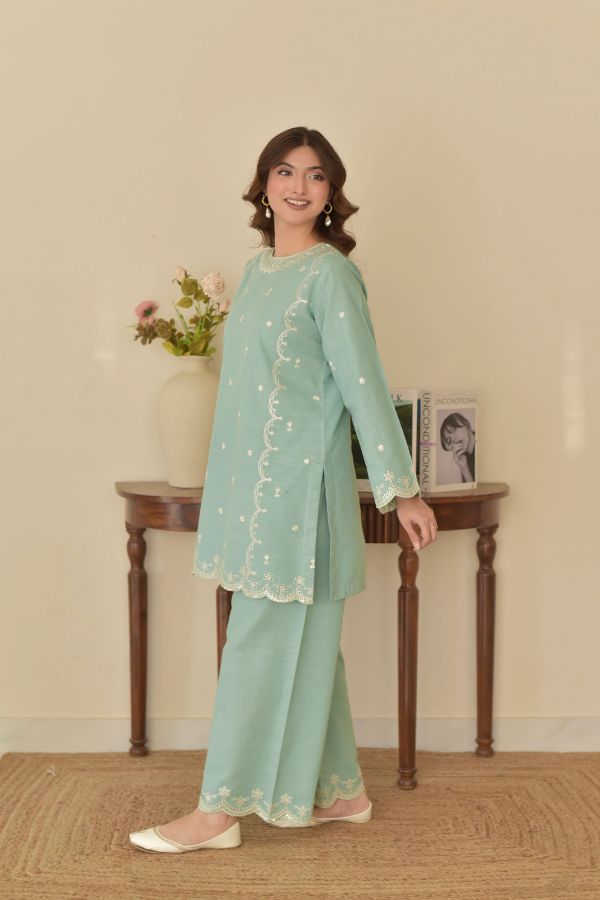 Woman wearing a light green traditional outfit with embroidery, standing in a room with a wooden table and decorative items.