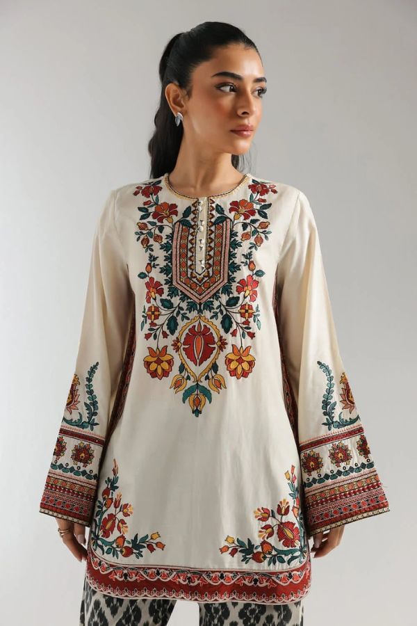 Woman wearing a cream-colored embroidered dress with colorful patterns on a plain background