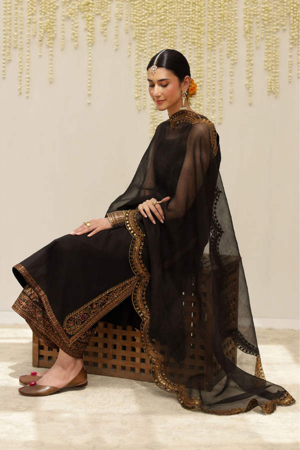 Woman in a black traditional outfit with gold embroidery sitting on a decorative chair.