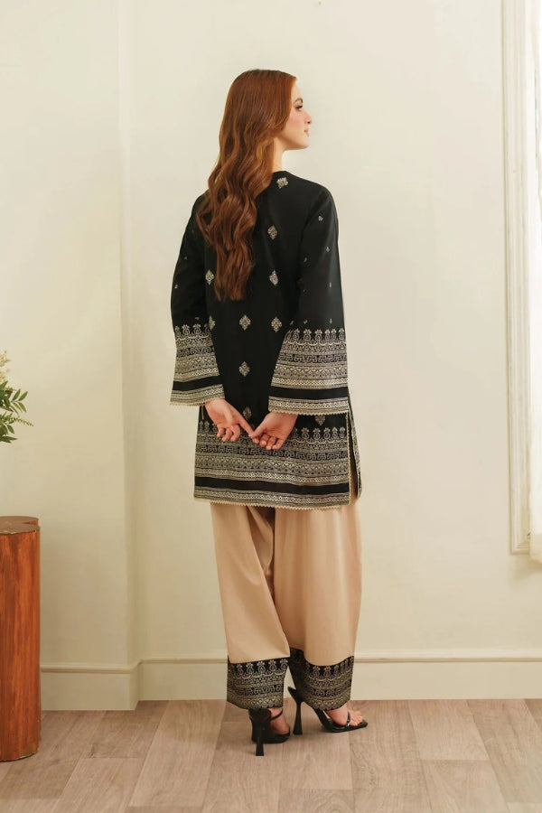 Woman wearing a black embroidered top and beige pants standing against a white wall.