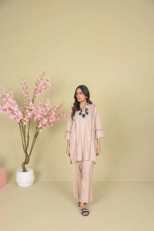 Woman in a beige outfit standing next to a plant with pink flowers against a beige wall.