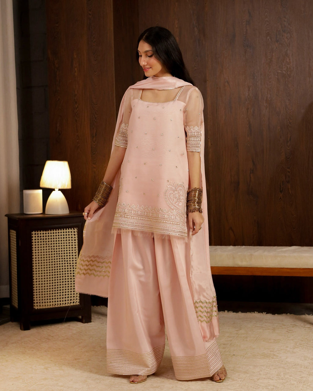 Woman in a pink traditional outfit standing in a room with a wooden wall and lamp.